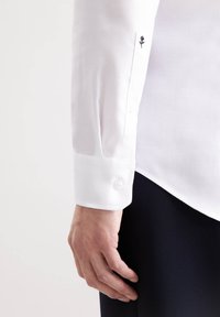Camicia bianca con texture a spina di pesce, polsino con un bottone e un piccolo ricamo a forma di rosa sul maniche. Mano visibile sotto.