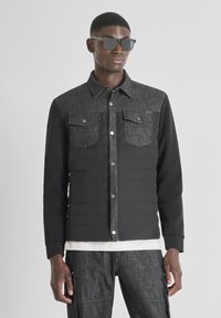 Giacca ibrida in denim nero e scuro con due tasche sul petto, bottoni a pressione e tessuto simile a un maglione sulle maniche. Indossata con pantaloni grigi.