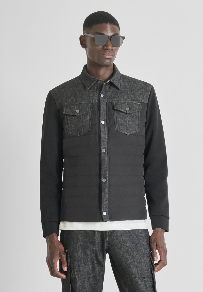 Giacca ibrida in denim nero e scuro con due tasche sul petto, bottoni a pressione e tessuto simile a un maglione sulle maniche. Indossata con pantaloni grigi.