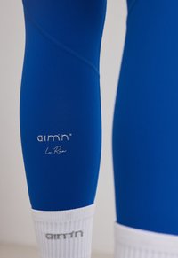 Personne portant des leggings de sport bleus avec le texte "aim'n Le Run" au-dessus de chaussettes blanches affichant le logo "aim'n".