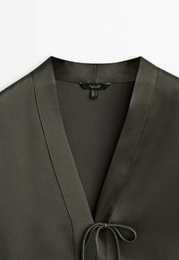 Blouse vert foncé avec un décolleté en V et une fine fermeture à nouer à l'avant, texture de tissu lisse et petite étiquette à l'intérieur du col.