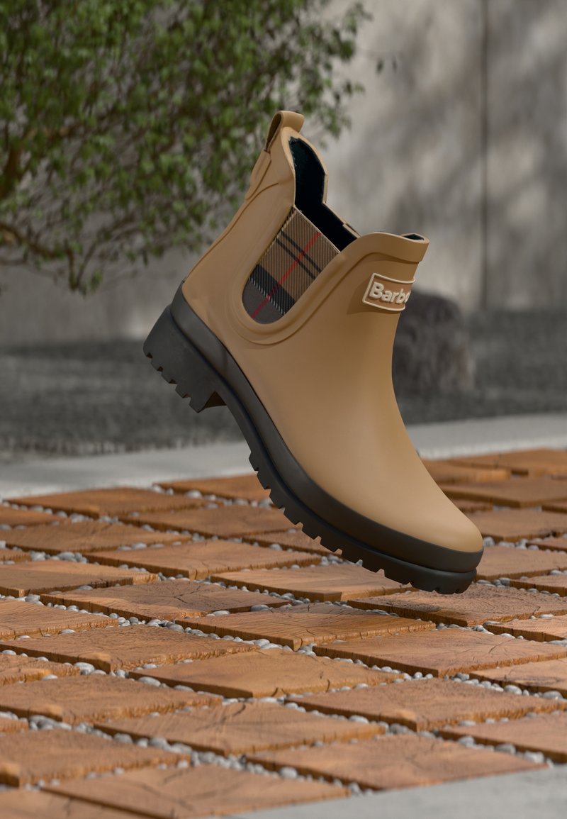 Καφέ λαστιχένια μπότα με μαύρη σόλα και καρό ελαστικό πλαϊνό πάνελ που αιωρείται πάνω από ξύλινο δάπεδο με πλακάκια σε εξωτερικό χώρο.