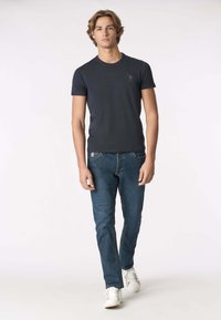 T-shirt a maniche corte di colore blu scuro con scollo rotondo e sottili dettagli con il logo, abbinata a jeans aderenti blu e scarpe da ginnastica bianche.