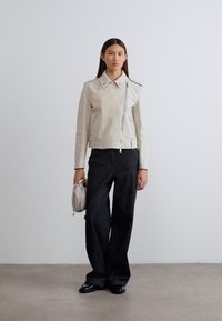 Ανοιχτό γκρι δερμάτινο μπουφάν μοτοσυκλέτας με φερμουάρ, μυτερό γιακά και μινιμαλιστικό σχέδιο. Συνδυασμένο με μαύρο παντελόνι φαρδιάς γραμμής και μικρό τσαντάκι.