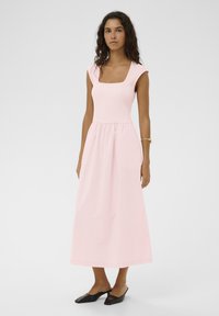 Rosa Kleid mit quadratischem Ausschnitt, tailliertem Oberteil und fließendem Rock. Hergestellt aus leichtem Stoff, mit kurzen Kappenärmeln. Schwarze Mules getragen.