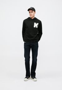 Felpa nera con un grafico geometrico bianco nella zona del petto, abbinata a jeans blu scuro e sneaker nere con suole bianche.