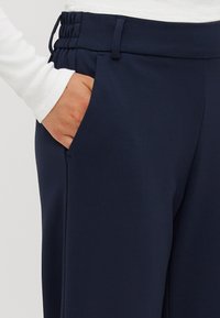 Námornické kalhoty s hladkým texture, disponující stahovací šňůrkou v pase, bočními kapsami a volným střihem. Zdůrazněné bílým rukávem v záběru.