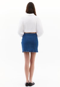 Vit, texturerad kort blus med puffärm; blå denim mini-kjol med bakfickor; svarta platta skor; visad bakifrån.