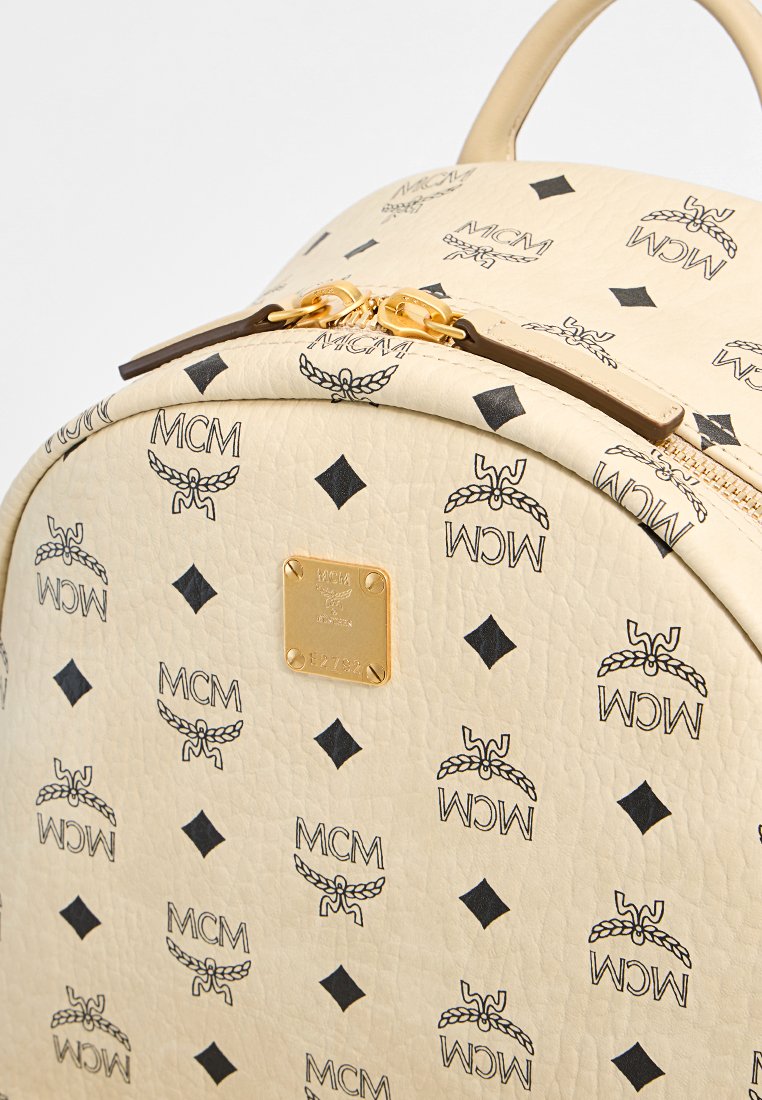 Beige Leder-Rucksack mit schwarzem MCM-Logo-Print, goldenen Hardware, runder Form und einer auffälligen Metallmarke.
