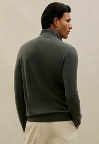 Tumši zaļš, mīksts adīts augstais kakls ar garām piedurknēm. Iezīmēts ar brīvu piegriezumu un ribotām piedurknēm un apakšmalu. Parādīts no aizmugures.