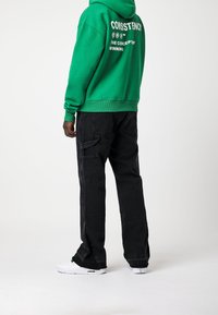 Felpa verde con testo bianco sulla schiena, abbinata a pantaloni cargo neri con tasche laterali e sneakers bianche con dettagli verdi.