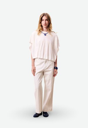 Femme debout portant un haut blanc ample, un pantalon large de couleur claire, un collier bleu, un bracelet et des chaussures plates foncées sur fond uni.