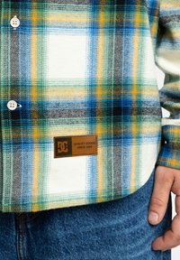 Koszula w kratę w białych, czarnych, zielonych, niebieskich i żółtych odcieniach; posiada guziki oraz metkę DC z napisem "Quality Goods Since 1994".
