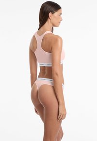 Lichtroze sportbeha en bijpassende string met witte elastische banden voorzien van de tekst "GUESS". Glad materiaal met een aansluitend ontwerp en racerstijl.