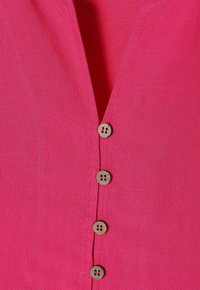 Camicia in lino rosa brillante a maniche corte con scollo a V profondo, caratterizzata da cinque bottoni rotondi in legno lungo la parte anteriore e una trama liscia.