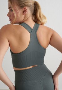 Sujeción deportiva de color verde oscuro con textura acanalada y diseño de espalda cruzada. Presenta un detalle recortado en la cintura, que complementa unos leggings de tiro alto.