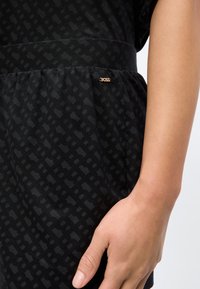 Κοντινό πλάνο του χεριού ενός ατόμου που ακουμπά κοντά στη μέση σε ένα μαύρο υφασμάτινο ένδυμα με μικρό χρυσό καρφίτσα λογότυπου "BOSS".