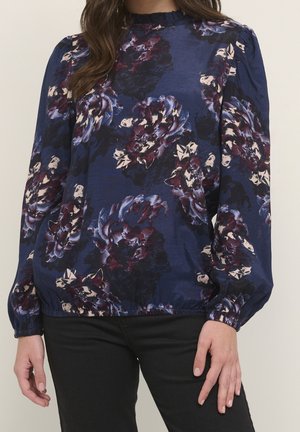 Femme portant un chemisier bleu marine avec de grands motifs floraux violets et blancs, des manches longues bouffantes et un col à volants, associé à un pantalon noir.