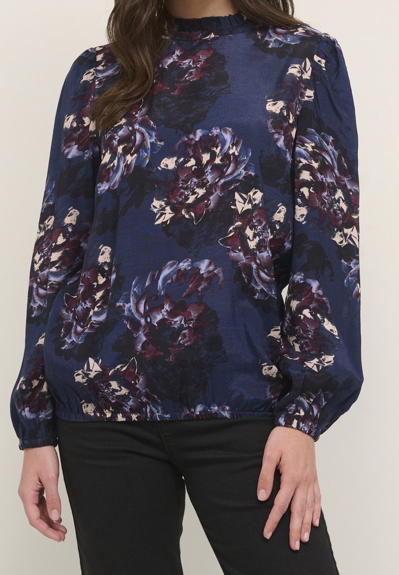 Femme portant un chemisier bleu marine avec de grands motifs floraux violets et blancs, des manches longues bouffantes et un col à volants, associé à un pantalon noir.