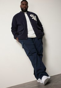 Marineblauwe fleece varsityjack met witte letters, gecombineerd met donkere cargobroek en witte sneakers, tegen een effen achtergrond.