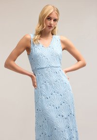 Rochie de culoare albastru deschis, cu decolteu în V, fără mâneci, și un model brodat complicat care seamănă cu evantaie. Material moale, texturat, cu decupaje detaliate.