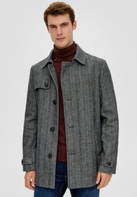 Grau-karierter Mantel mit Kragen, Knopfverschluss und seitlichen Taschen. Getragen über einem weinroten Rollkragenpullover und blauen Jeans, im lässigen Schnitt.