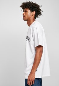 Camiseta de algodón blanca con mangas cortas, que presenta texto negro en la parte frontal. El diseño tiene un corte holgado y una abertura en el costado del dobladillo.