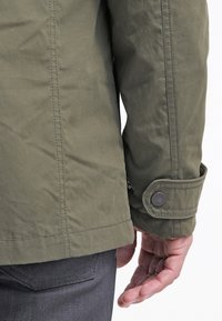 Olive green jacket with a smooth fabric texture, featuring a buttoned cuff and visible stitching details. The design is standard-fit with a straight cut.