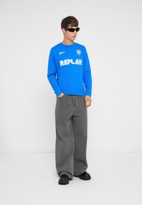 Camisa azul de mangas longas com "REPLAY" impresso em branco, combinada com calças largas cinzas e sapatos slip-on pretos.