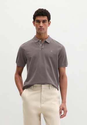 Mann mit lockigem Haar, der ein graues Poloshirt und beige Hosen trägt, steht mit einer Hand in der Tasche vor einem schlichten Hintergrund.