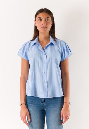 Giovane donna con lunghi capelli castani che indossa una camicia a righe a maniche corte azzurro chiaro e jeans blu, in piedi contro uno sfondo bianco semplice.