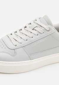 Baskets en cuir gris Calvin Klein avec texture perforée, lacets plats couleur crème et semelle en caoutchouc. Présentent un logo sur le côté et un design minimaliste.