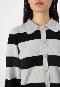 Harmaa ja musta raidoitettu neuletakki, jossa on kauluksellinen kaula-aukko. Takissa on teksturoitu kaulus ja nappikiinnitys edessä.