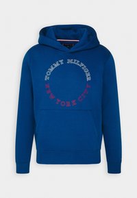 Modrá mikina s kapucí vyrobená z měkké směsi bavlny. Obsahuje kulaté logo v bílé a červené barvě, přední klokaní kapsu a žebrované manžety.