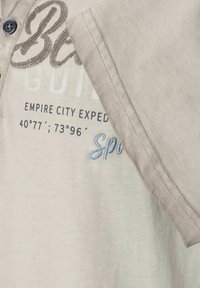 Jasno beżowa bawełniana koszula z zapinanym dekoltem, haftowanym napisem w brązowym i niebieskim kolorze oraz gładką fakturą.