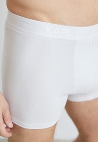 Bílé boxerky vyrobené z měkké tkaniny, s hladkým elastickým páskem, na kterém je vystouplý nápis "KARL LAGERFELD" v bílé barvě.