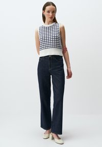 Biały dzianinowy top w stylu crop z granatowym wzorem w kratę, zestawiony z ciemnoniebieskimi spodniami na wysokim stanie. Modelka nosi beżowe buty na słupku.
