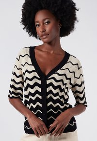 Strickjacke mit welligem schwarz-weißen Muster, kurzen Ärmeln, tiefem V-Ausschnitt und vorderem Knopfverschluss.