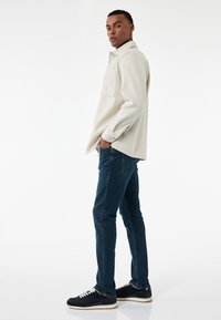 Ljust beige knäppskjorta, blå jeans och svarta sneakers. Skjortan har en bröstficka och långa ärmar. Slät textur.