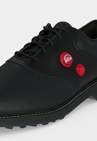 Chaussure en cuir noir avec lacets noirs et deux boutons de pompe rouges sur le côté, un étiqueté "The Pump" avec une semelle texturée.