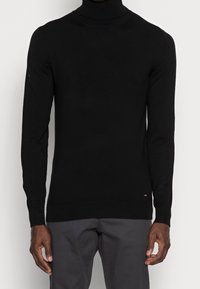 Muž v přiléhavém černém roláku a tmavě šedých kalhotách, stojící s uvolněnýma rukama podél těla.