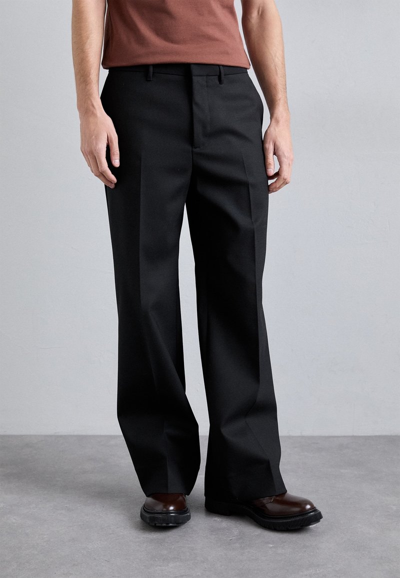 Homme portant un pantalon large noir associé à des chaussures en cuir marron et un haut d'un rouge brun foncé, debout sur un sol gris.