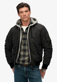 Sort bomberjakke med gråt hættelining, båret over en ternet flannelskjorte. Har en lynlåslomme på ærmet og ribstrikkede ærmekanter.