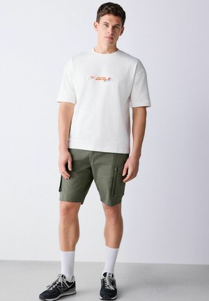 Junger Mann steht und trägt ein weißes T-Shirt mit Grafik, olivgrüne Cargoshorts, weiße Sportsocken und schwarz-weiße Turnschuhe.