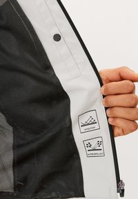 Hand, die eine offene Jacke hält, die mit schwarzem Netzfutter, Reißverschluss, Druckknopf und den Innenlabels "Wind Stop" und "Wasserabweisend" versehen ist.