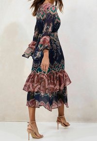 Proudící tmavě modré šaty s paisley vzory v růžové a zelené, volánkový lem, dlouhé průhledné rukávy a v páru s nahými vysokými podpatky.