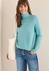 Hellblauer Rollkragenpullover aus weichem Strickmaterial, mit gerippten Akzenten am Kragen und an den Ärmeln, kombiniert mit Jeans und einer weißen Tasche.