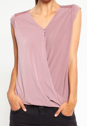 Bez rękawów bluzka w kolorze mauve z koronkowymi detalami na ramionach i kopertowym przodem, lekko wsunięta w czarne spodnie, z rękami w kieszeniach.