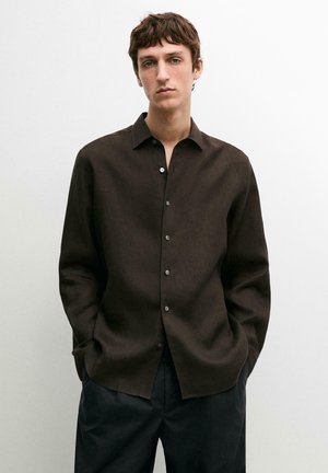 Junger Mann steht mit den Händen in den Taschen, trägt ein dunkelbraunes Hemd mit Knöpfen und schwarze Hose vor einem einfarbigen Hintergrund.