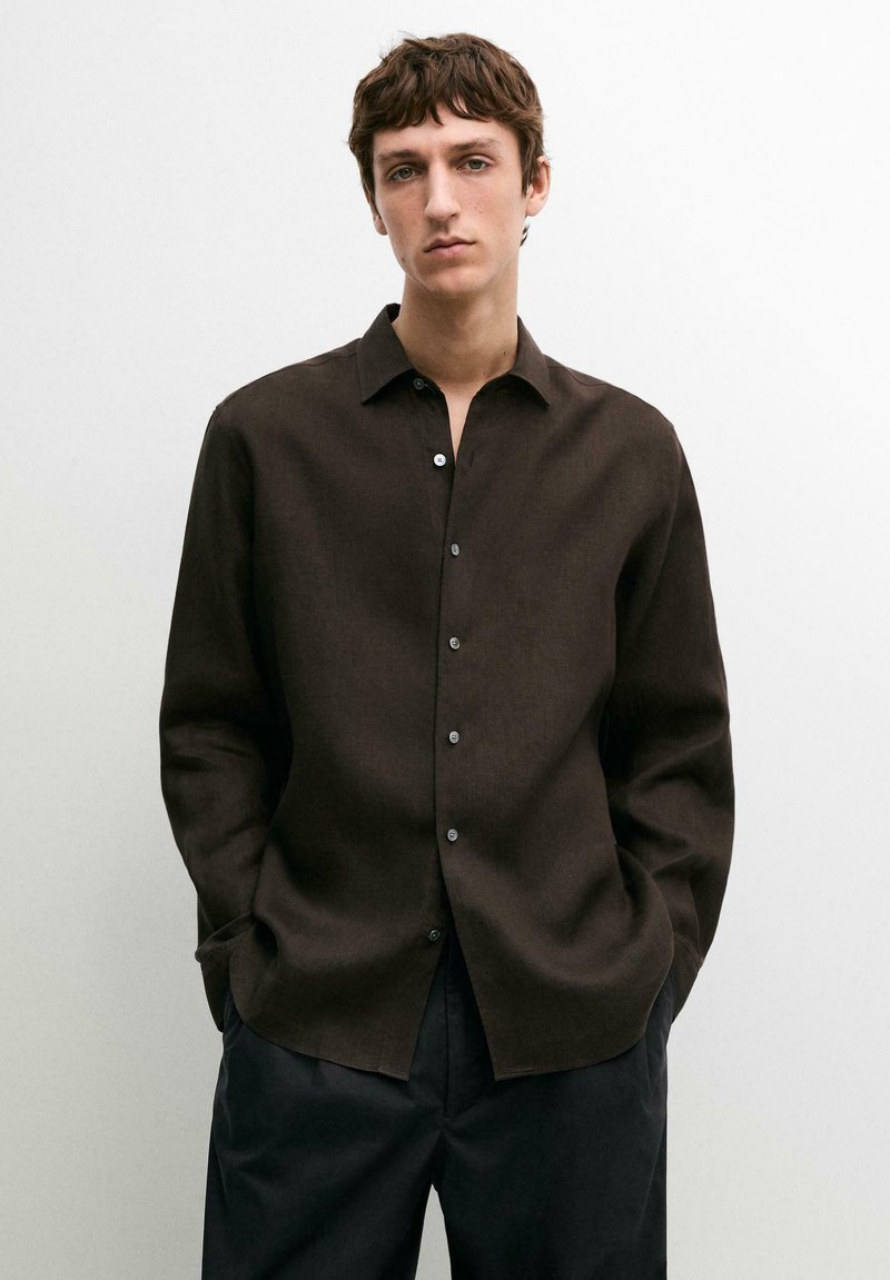 Giovane uomo in piedi con le mani nelle tasche, indossa una camicia marrone scuro con bottoni e pantaloni neri su uno sfondo semplice.